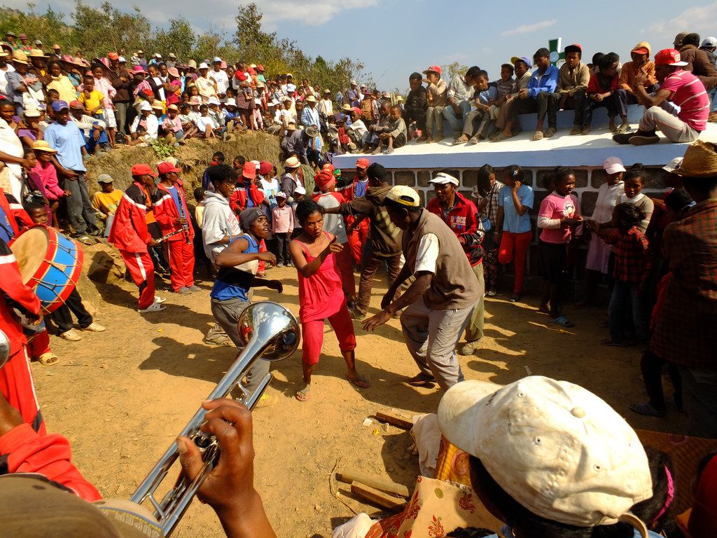 Kolorowy obrazek przedstawiający uczestników ceremonii famadihana. Widać bliskich zmarłego, którzy tańczą, grają na trąbkach, śpiewają tradycyjne pieśni ku jego czczi. Na środku wielkiego koła tańczy dwóch mężczyzn, a wokół nich zebrana jest liczna grupa ludzi.