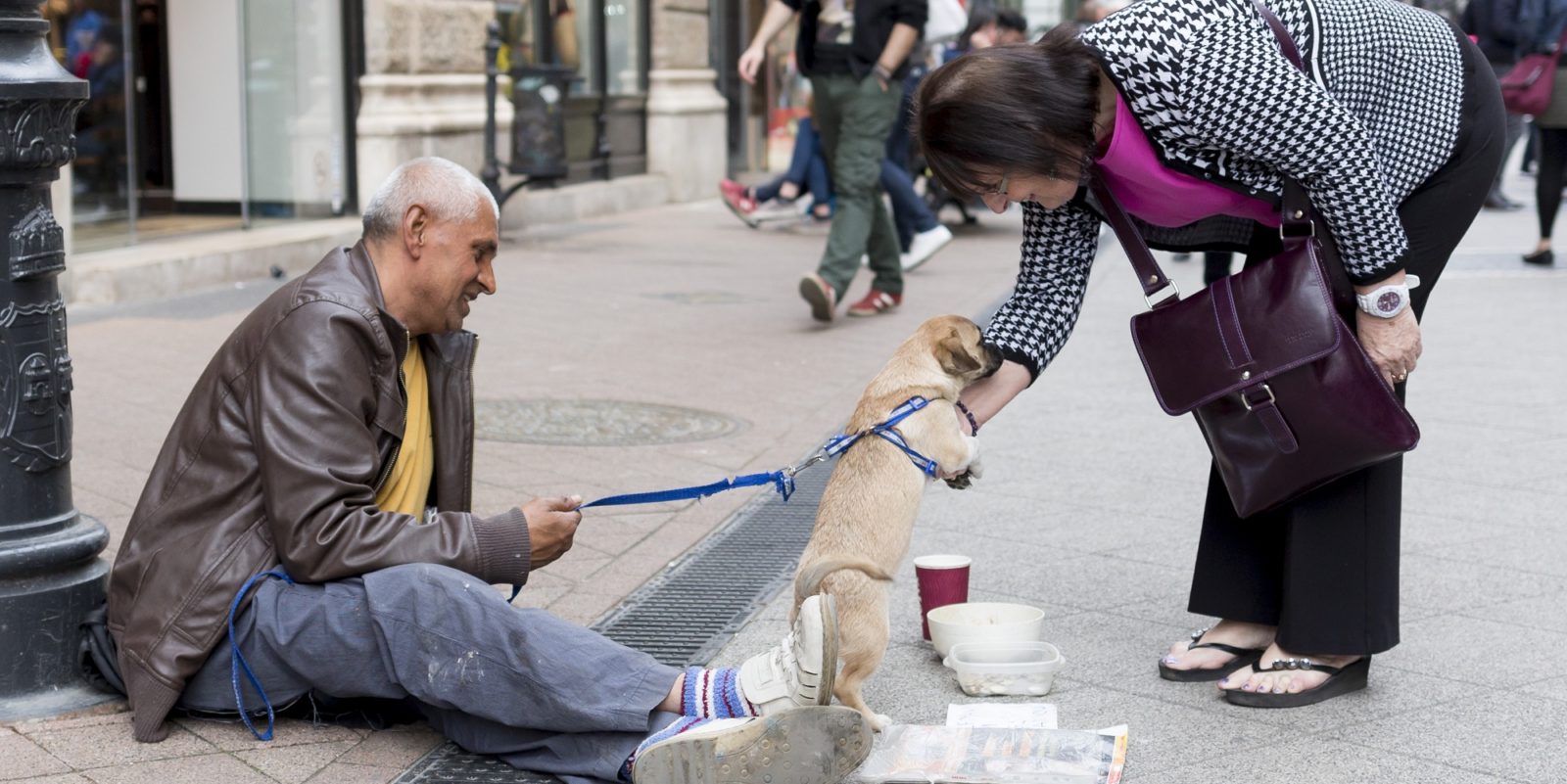 Zdjecie bezdomnego z psem, przy ktorych zatrzymala sie kobieta. Ona bawi się z psem, on siedzi na ulicy. Pies szczeniaczek, jest trzymany na smycyz przez bezdomnego