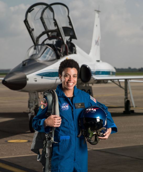 na zdjęciu jest kobieta w kostiumie astronauty, trzyma w ręku kask a za nia jest mały samolot