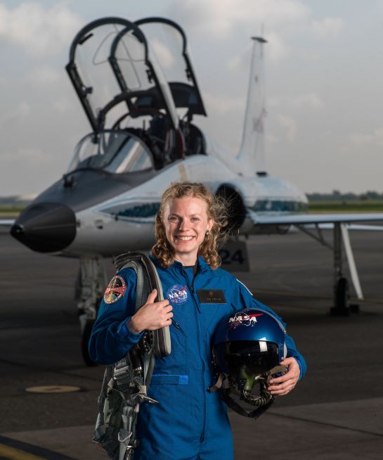 na zdjęciu jest kobieta w kostiumie astronauty, trzyma w ręku kask a za nia jest mały samolot