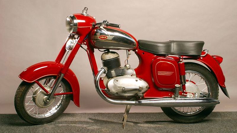 Na zdjęciu widoczny jest dawny motocykl Jawa. Stoi na jasnym tle. Jest koloru czerwonego