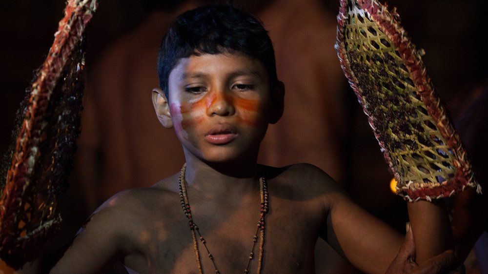 Fotografia młodego chłopca z plemienia Satere Mawe, który przechodzi proces stawania się mężczyzną. W tym celu przez 10 minut ma na rękach specjalnie tkane rękawice, które wcześniej wypełnioną mrowkami buna. Stoi spokojnie z rękami podniesionymi do góry, na twarzy ma wymalowane specjalne wzory.