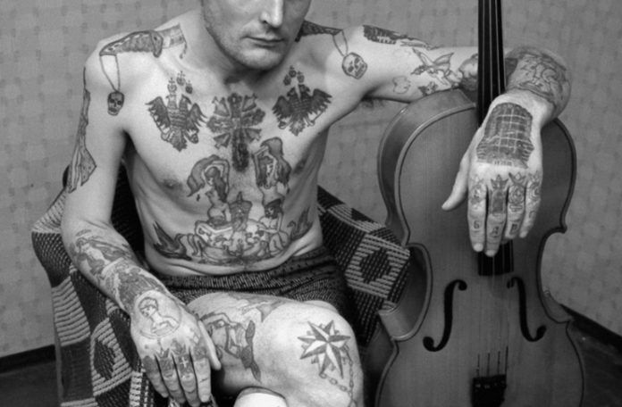 Zdjęcie rozebranego do bielizny mężczyzny, który jest cały w radzieckim tatuażach. Siedzi na fotelu, trzyma nogę na nodze a lewą rękę opiera o skrzypcę.