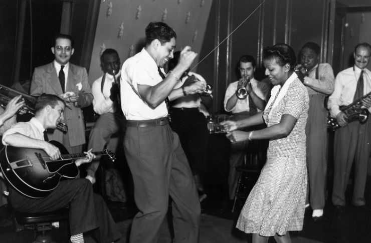 Czarno-białe zdjęcie tańczącej pary. Do okoła nich widać grających na żywych instrumentach muzyków. Kobieta ubrana w wzorzystą sukienkę, mężczyzna w spodnie od garnituru i biała koszulę.