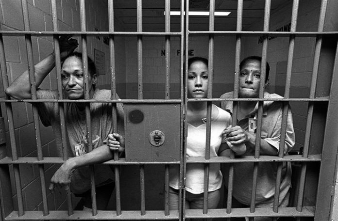 Czarno białe zdjęcie przedstawiające trzy kobiety znajdujace się za kratami więzienia. Każda z nich stoi przy kracie i lekko wystawia głowę