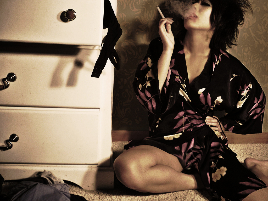 Ciemnowłosa kobieta siedząca na podłodze, obok komody, paląca papierosa i wydmuchująca w górę dym.