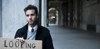 Schludnie ubrany młody człowiek siedzi na pustej ulicy z tabliczką na której napisał ' looking for a job'.