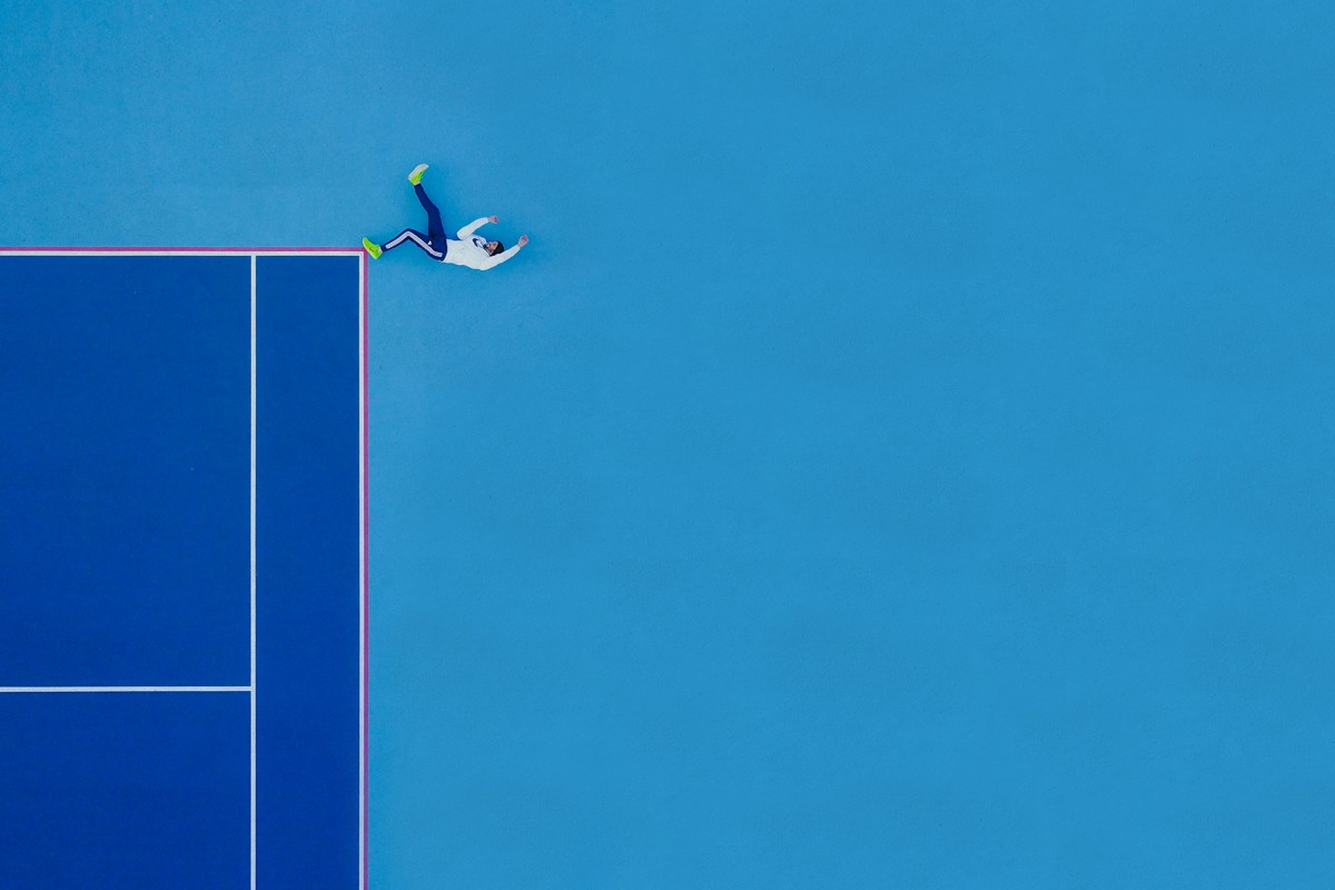 Zdjecie z lotu ptaka, ktore przedstawia boczną częśc boiska od tenisa i mezczyzne, ktory lezy na jego rogu. Wszystko razem tworzy jakby człowiek spadał z wysokosci. Boisko i tło dookoła boiska są niebieskie, mezczyzna ma na sobie białą bluzę
