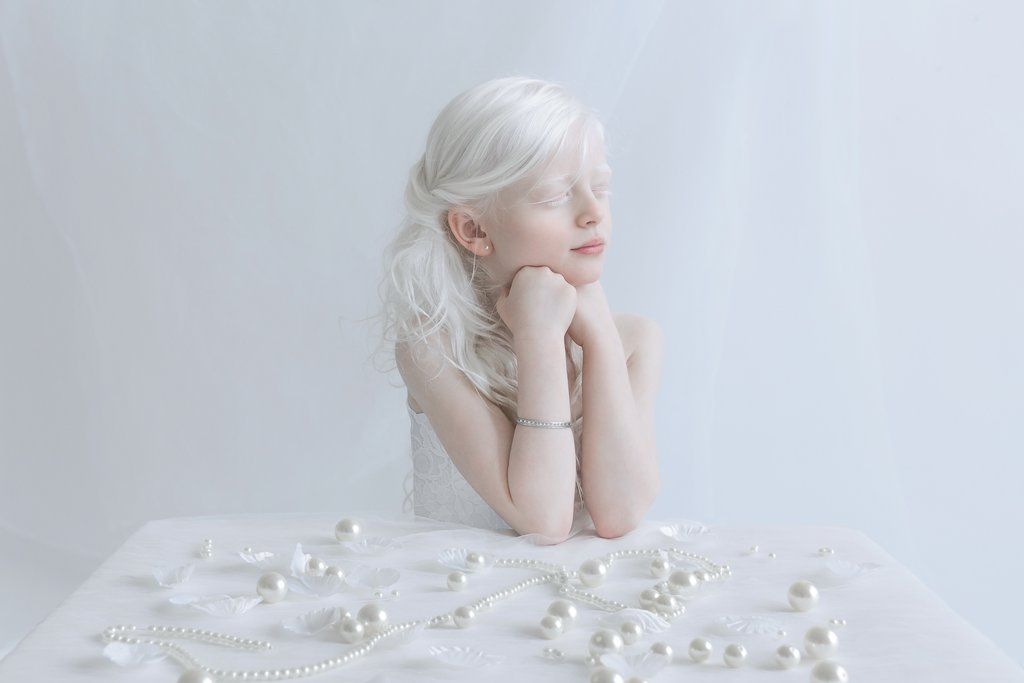 Fotografia kolorowa utrzymana w białych odcienaich. Na zdjęciu widać dziewczynkę-albinos siedzącą przy białym stole, opiera ona ręce o blat, podpierają również brodę. Ma zamknięte oczy. Na blacie rozrzucone są białe perły.