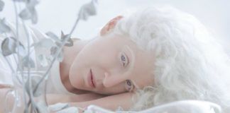 Fotografia kolorowa utrzymana w białych barwach. Na zdjęciu widać kobietę-albinosa, z błekitnymi oczami leżącą wśród białych kwiatów.