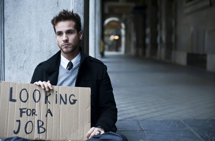 Schludnie ubrany młody człowiek siedzi na pustej ulicy z tabliczką na której napisał ' looking for a job'.