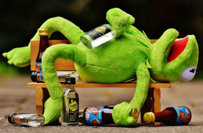 Zaba kermit leży na ławce trzymając w dłoni butelkę alkoholu, a wokół niego porozrzucane są kolejne butelki alkoholu