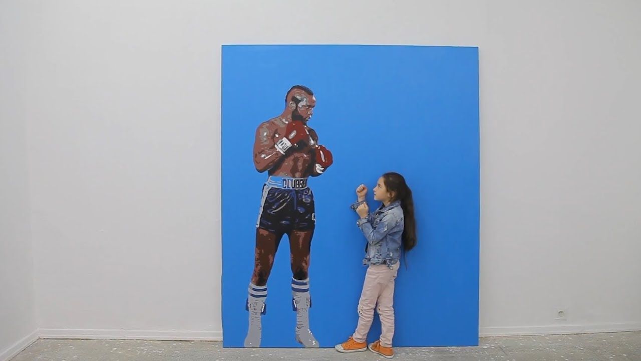 Obraz boksera czekajacego na walkę, uzupełniony przez dwa razy mniejsza dziewczynkę trzymająca gardę. Czarnoskory bokser namalowany jest na niebieskim tle. Dziewczynka ma na sobie jeansowa kurtkę, jasne spodnie i czarne włosy.