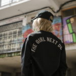 Dziewczyna na tle graffiti odwrócona tyłem w czarnej kurtce z napisem The Girl Next Door