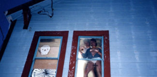 Zdjecie robione nocą. Widać na nim parę uprawiająca seks w oknie. Kobieta przylega do szyby. Pomiędzy nogami trzyma krzyż. Mężczyzna jest za nią. Widać białe ściany domu.