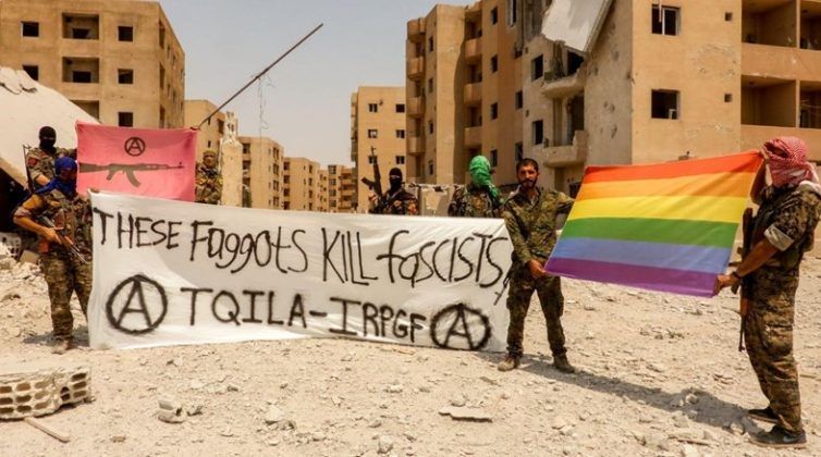 Zdjecie żołnierzy-ochotników, ktorzy trzymaja flagi srodowiska LQBT oraz transparent z haslem these faggots kil facists. W tle widac miasteczko Syrii, ktore jest dotkniete wojną i zniszczeniem.