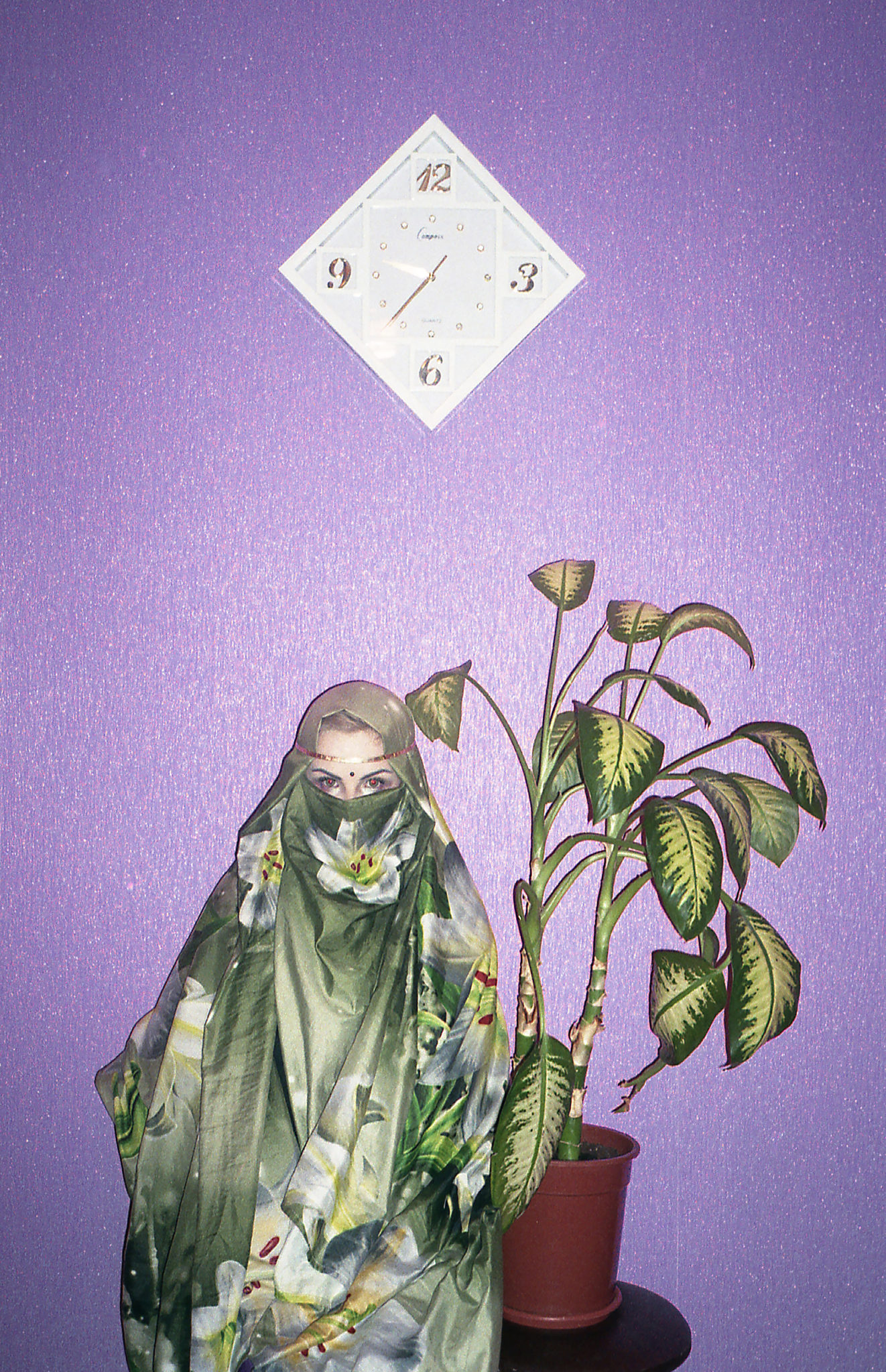 Młoda kobieta stoi ubrana w burce z kwiatami. Przy niej stoi roślina w doniczce, a nad nią znajduję się zegar w kształcie kwadratu.