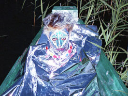 Młody mężczyzna cały pomalowany na twarzy siedzi w folii aluminiowej na łódce pośród trzcin.