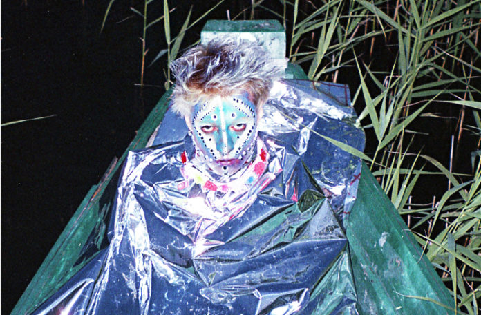 Młody mężczyzna cały pomalowany na twarzy siedzi w folii aluminiowej na łódce pośród trzcin.