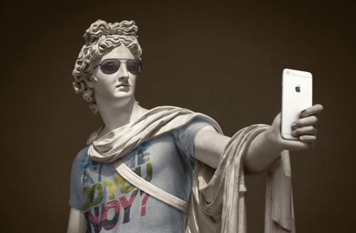 Klasyczna rzeźba ubrana w koszulkę, szalik, okulary, trzymająca w ręku iPhone'a
