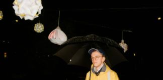 Biały chłopiec w żółtej kurtce z czapką, trzyma parasolkę podczas mocnej ulewy na tle lampionów.