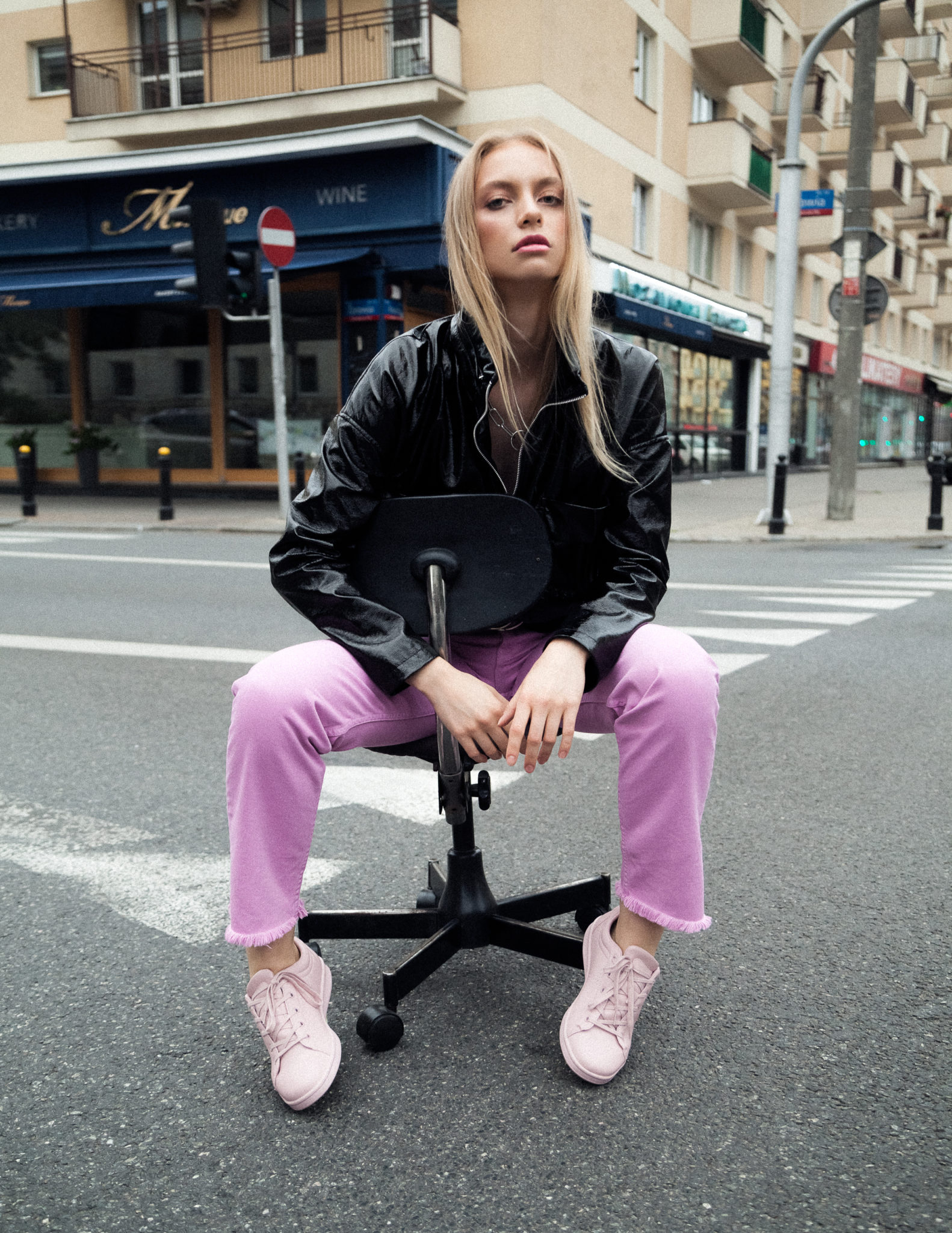 Dziewczyna w różowych spodniach i czarnej kurtce siedzi na środku ulicy na krześle z kółkami