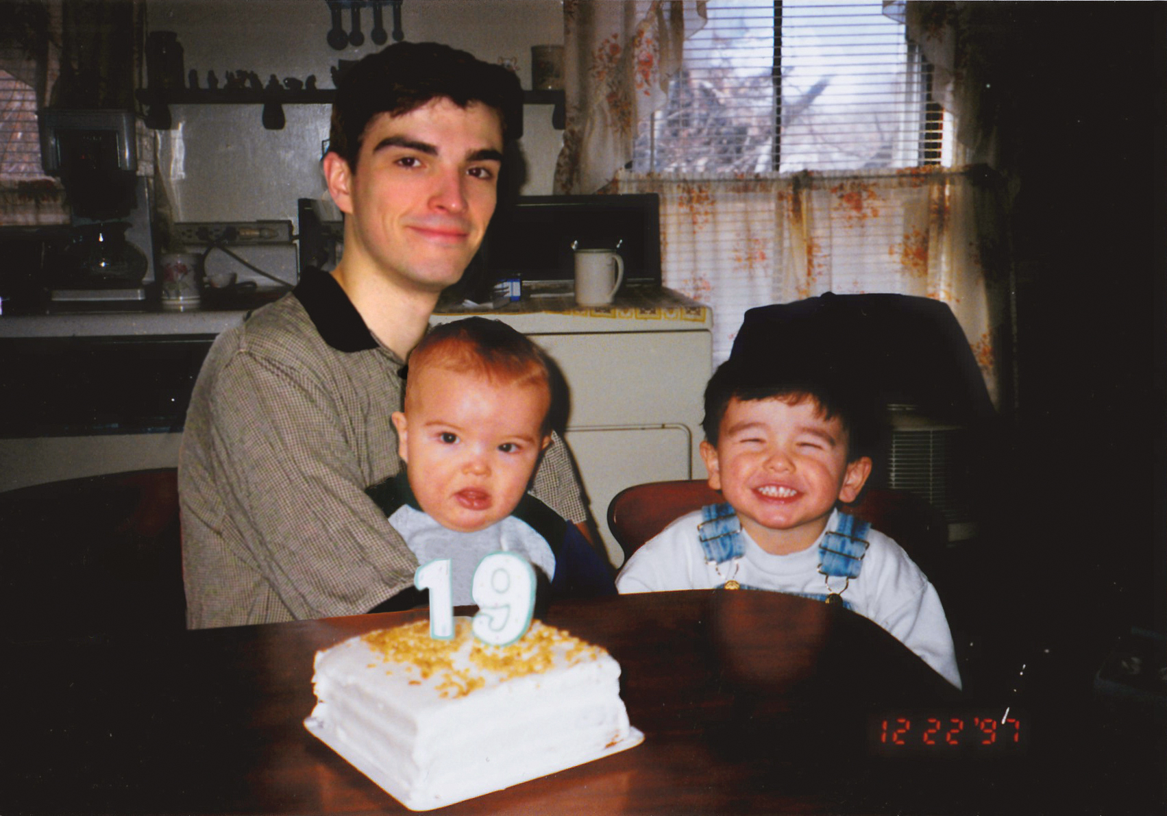 Mężczyzna siedzi na krześle z dzieckiem na kolanach. Obok po prawej starsze śmiejące się dziecko. Przed nimi na stole tort ze świeczkami 1 i 9