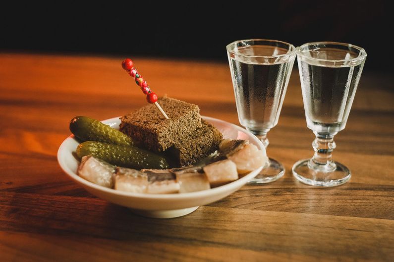 Dwa kieliszki wódki, a obok na talerzu ogórki i chleb z wbitą wykałaczką. Wszystko stoi na stole