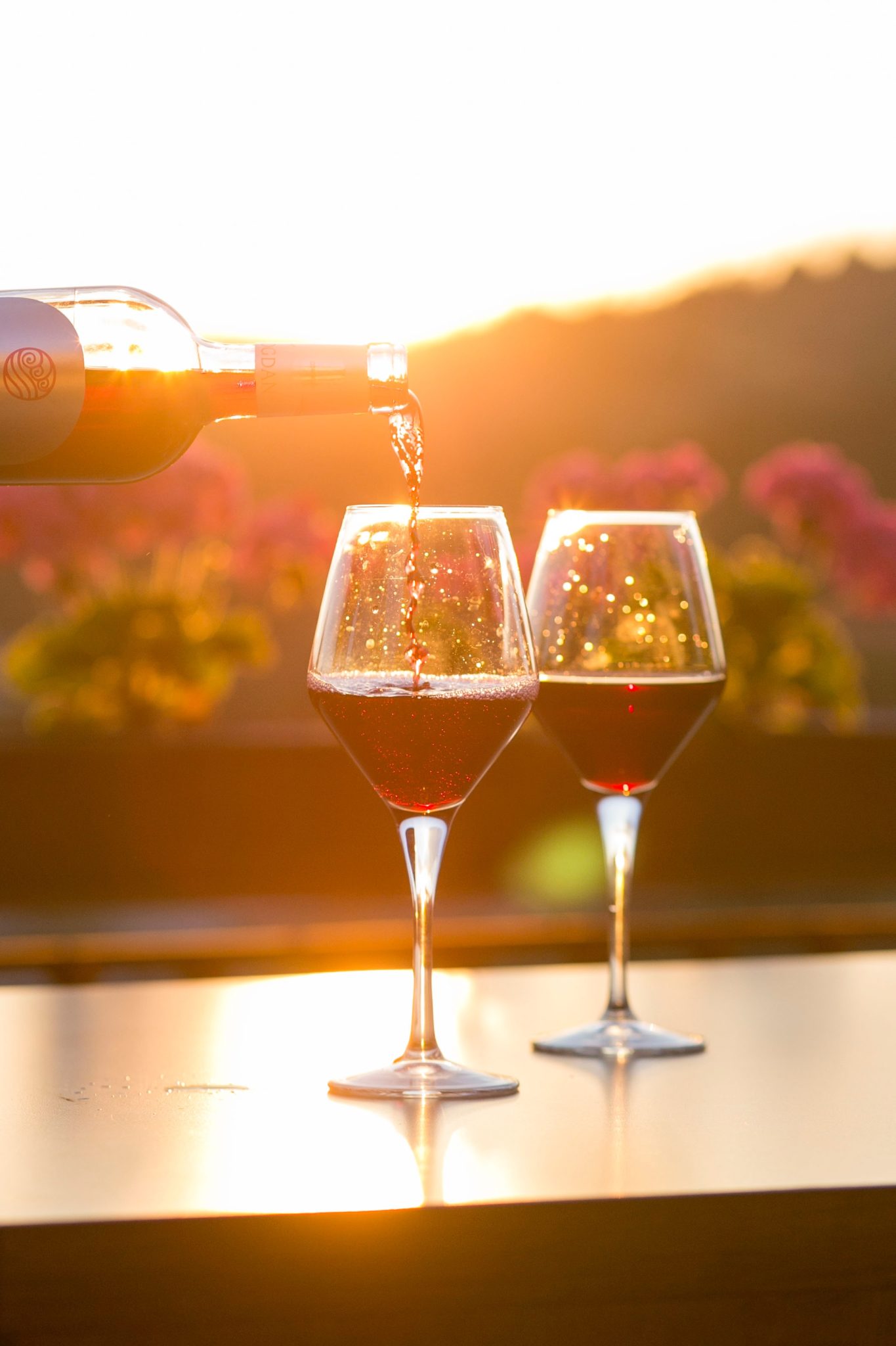 Dwie lampki czerwonego wina stojące na stole, a w tle ogród o zachodzie słońca