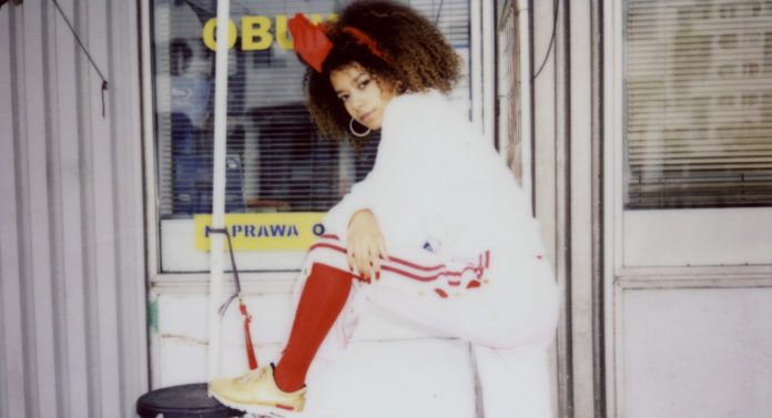 Analogowe zdjęcie przedstawiające dziewczynę z afro na głowie ubraną w biały dres, czerwone skarpetki i złote buty