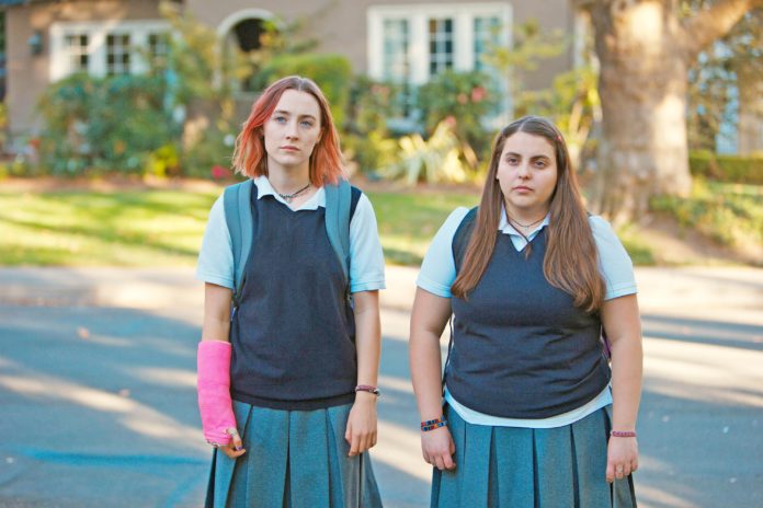 Dwie, stojące na ulicy dziewczynki w szkolnych uniformach. Dziewczyna z lewej ma różowy gips i czerwone włosy.