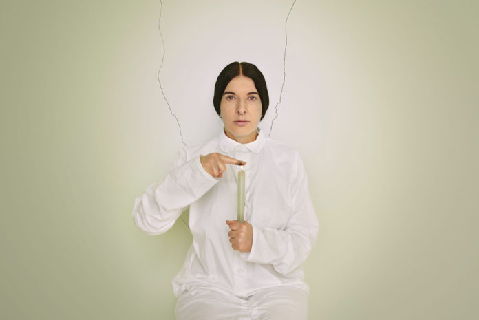 Kobieta w czarnych, związanych włosach w białym kombinezonie na tle białej ściany, trzyma w ręku białą, palącą się świecę, palec drugiej ręki przystawia do płomienia.