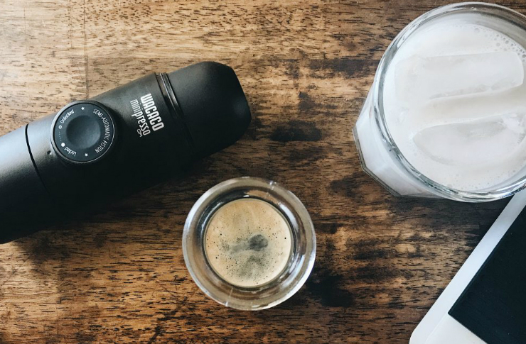 Przenośny ekspres do kawy leżący na stole, obok szklanka z espresso, szklanka z mlekiem i telefon