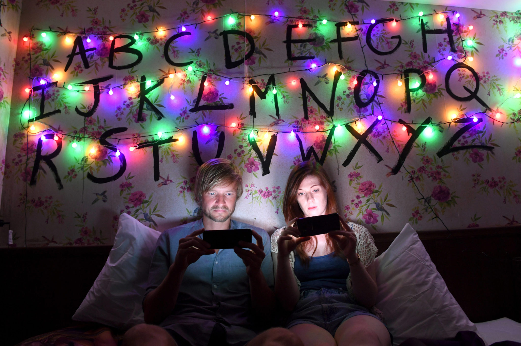 Ciemny pokój oświetlony kolorowymi lampkami, na ścianie wypisany alfabet. Na łózku chłopak i dziewczyna patrzą w świecące telefony komórkowe.