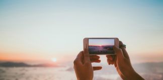 Dłonie trzymające telefon komórkowy, który robi zdjęcie zachodowi słońca nad morzem.