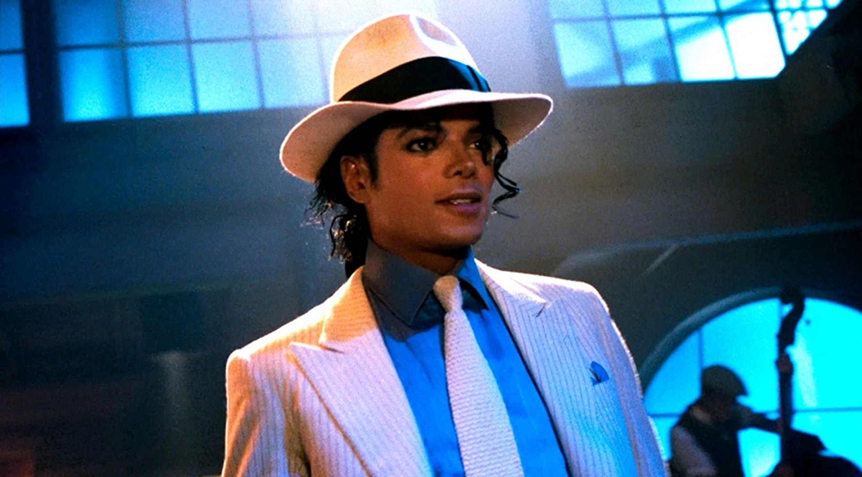 Ciemnoskóry mężczyzna z czarnymi lokami w białym kapeluszu, białym krawacie, niebieskiej koszuli i białej marynarce.