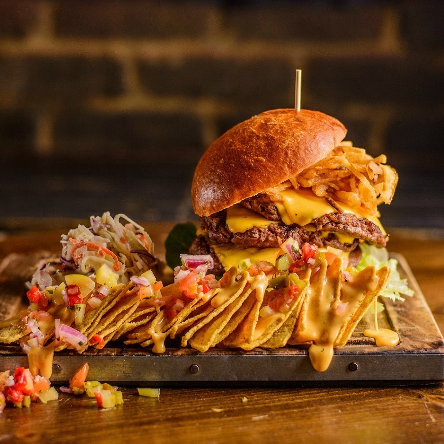 Kolorowe zdjęcie jedzenia fast food - burger, ociekające sosem nachosy i warzywa podane na drewnianej desce na tle ściany z cegieł
