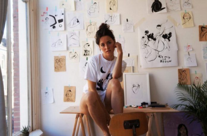 Na zdjeciu widzimy mloda kobiete siedzaca w swojej pracowni artystycznej pelnej szkicow i rysunkow