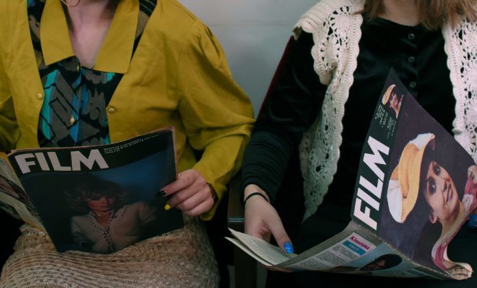 dwie dziewczyny czytające magazyny filmowe, jedna w żółtej marynarce, druga w czarnym swetrze, kadr ucina nogi i głowy