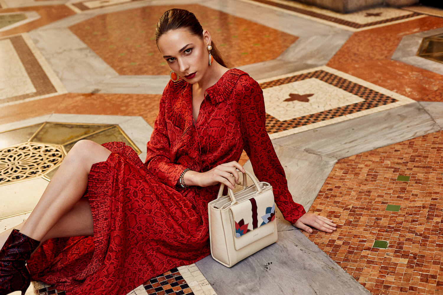 Na zdjeciu widzimy mloda kobieta ubrana w czerwona sukienke z bezowa wzorzysta torebka siedzi ona na kolorowym okazalym dywanie
