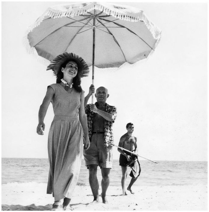 Kobieta spacerująca po plazy, za nia mezczyzna z parasolem