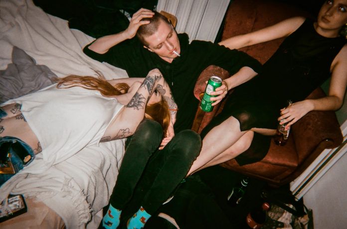 Dziewczyna leżąca na łóżku, chłopak z butelką piwa i dziewczyna na fotelu
