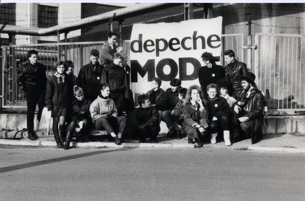 czarno-białe zdjęcie przedstawiające grupę ludzi ubranych w czarne ramoneski, którzy stoją na tle baneru z napisem Depeche Mode