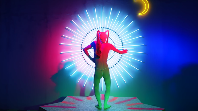 Kadr z teledysku Aishy Devi - Mazda przedstawiający postać artystki stojącej tyłem na tle emanującej energią aureolidowej elektroniki