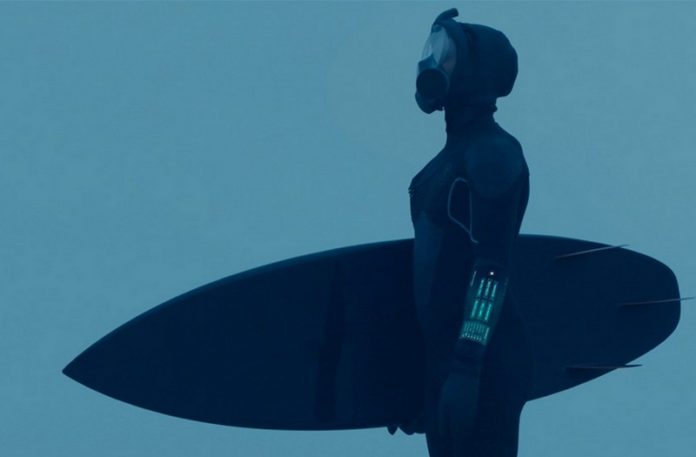 Postać w ciemnym stroju pływackim, zakrywającym głowę maską, trzyma deskę do surfingu
