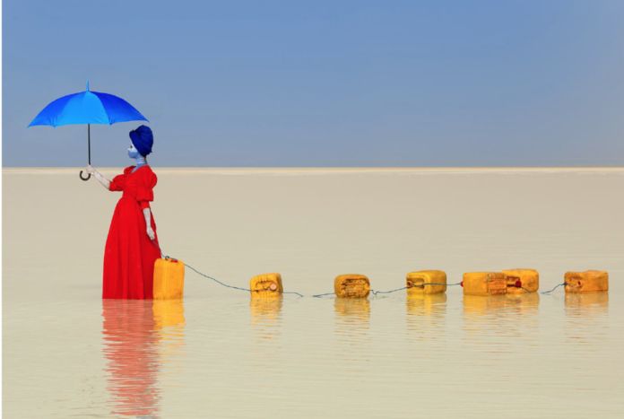 Zdjęcie przedstawia kobietę ubraną w czerwoną suknię trzymającą błękitny parasol na tle błękitnego nieba. Idzie ona przez jezioro, ciągnąc za sobą żółte kanistry na wodę