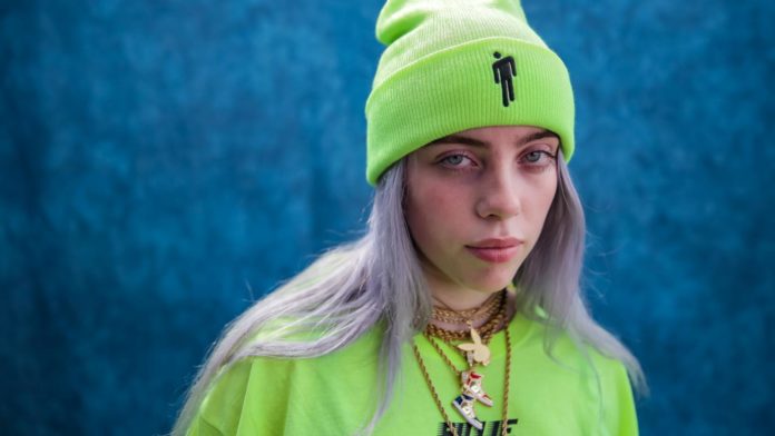 Zdjęcie przedstawia młodą artystkę BBillie Eilish, w jaskrawo zielonej czapce i bluzce w tym samym kolorze. Na szyi ma wiele, złotych wisiorów